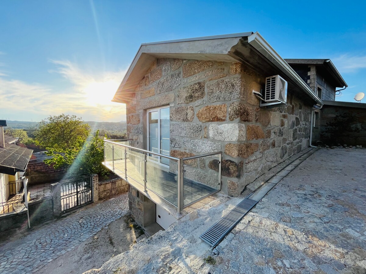 The exterior of the stone house is depicted, showcasing a modern balcony with glass railing. Natural light from the setting sun enhances the warm tones of the stone, while the surrounding landscape includes trees and distant views. An air conditioning unit is visible on the side.