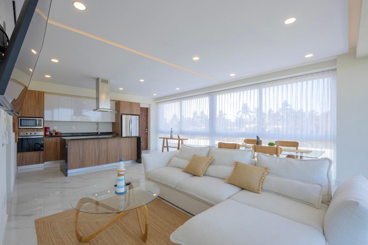 A modern living space is showcased, featuring a comfortable sectional sofa adorned with decorative cushions. Large windows provide ample natural light, with sheer curtains allowing for privacy. A dining table and stylish glass coffee table complement the kitchen area, creating an inviting atmosphere for relaxation.