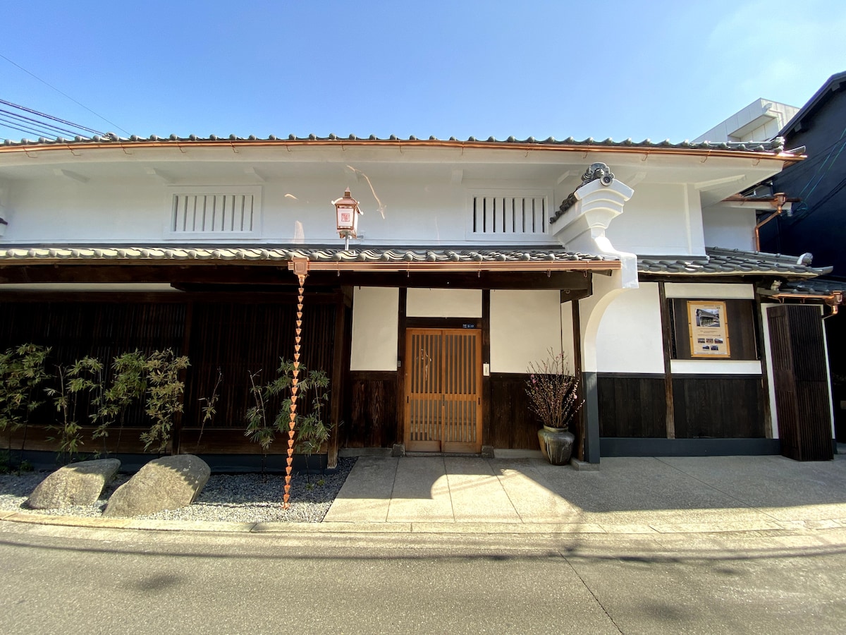 The exterior of a traditional Japanese building is shown, featuring a wooden entrance door and stone pathway. The structure is characterized by white walls, a tiled roof, and decorative lanterns. Greenery is present in the form of small trees and plants lining the pathway.