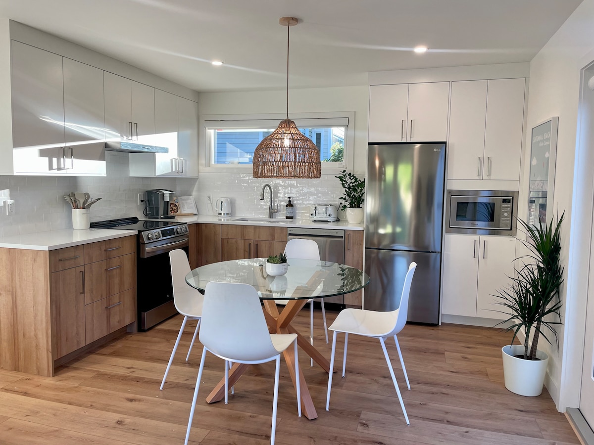A modern kitchen area features light wood cabinetry contrasted by sleek white cabinetry. A round glass table with light wood legs is surrounded by four white chairs. A large metal refrigerator and microwave are present, while potted plants add a touch of greenery.