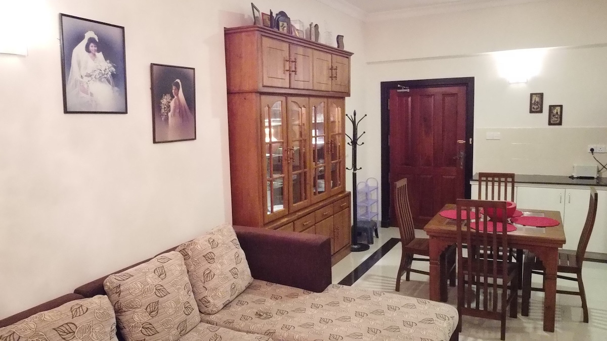 A cozy living area is presented with a plush beige sofa adorned with subtle patterns. A wooden dining table is set for four, complemented by matching chairs. On the wall, framed photographs add personal touches, while a wooden cabinet displays decorative items.