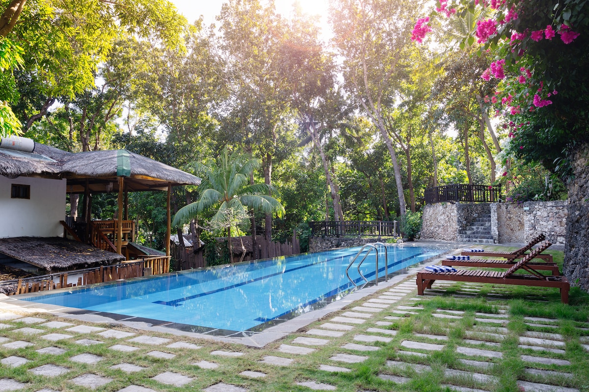 A serene swimming pool is surrounded by lush greenery and flowering plants. Sun loungers are positioned on a manicured lawn, while a wooden gazebo is visible nearby, providing shaded seating. The area is bathed in warm sunlight, enhancing the peaceful atmosphere.