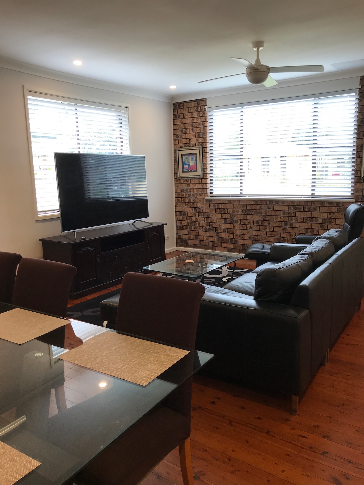 A cozy lounge area is presented with a large flat-screen television mounted on the wall. Comfortable seating includes a black leather sofa, complemented by a glass coffee table. Bright light filters in through window blinds, enhancing the warm wooden flooring.