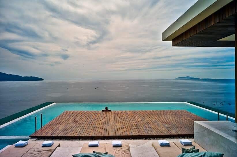 An infinity pool extends toward the horizon, merging with the ocean. Wooden decking surrounds the pool, with lounge chairs arranged neatly. The sky is softly clouded, creating a serene atmosphere. The distant hills are visible across the calm water.