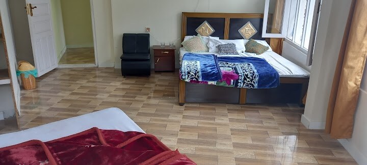 A spacious room is presented with a cozy bed adorned in vibrant blankets. A small seating area includes a dark armchair beside a wooden nightstand. Natural light enters through open windows, highlighting the clean wooden floor.