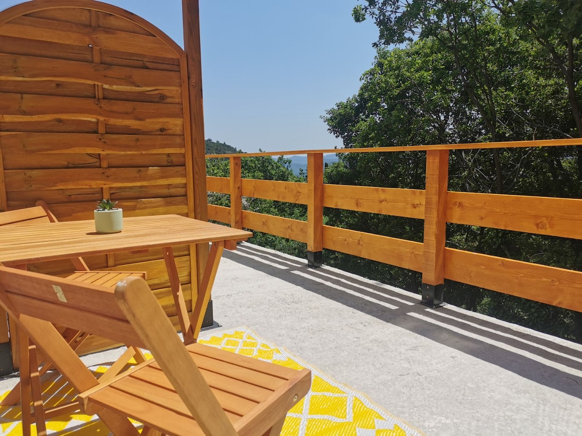 An outdoor terrace features wooden railings and furniture, including a table and chairs. A patterned yellow rug lies on the flooring, enhancing the relaxing space. Lush green trees are visible in the backdrop, with a glimpse of the distant landscape under clear blue skies.