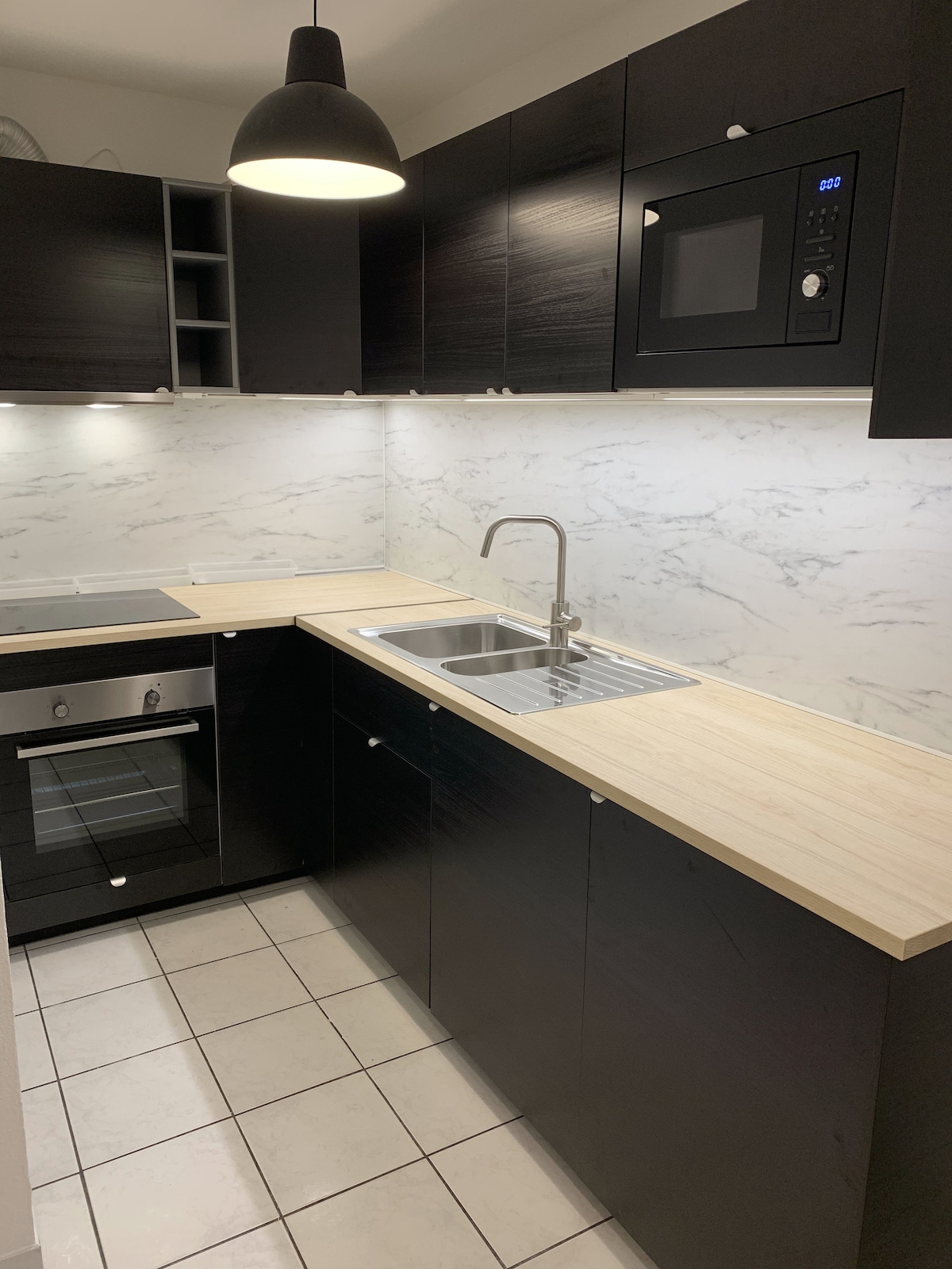 The kitchen is equipped with dark cabinetry and a light wood countertop. A modern sink is positioned beneath wall-mounted cabinets. Appliances include a built-in microwave, an oven, and a stovetop, all integrated into the counter design. A pendant light hangs above, illuminating the space.