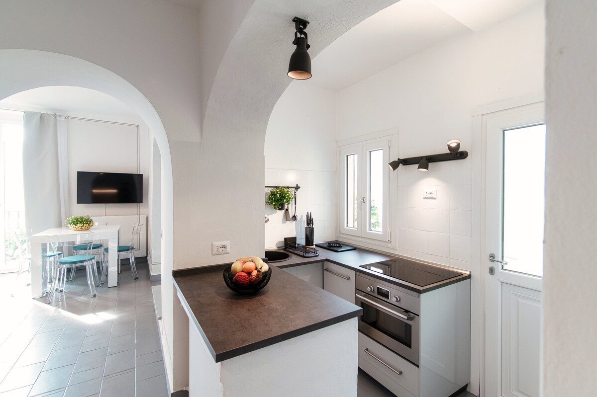 The kitchen features modern appliances and a dark countertop, complemented by white cabinetry and a clean aesthetic. An archway leads to the dining area, which includes a table surrounded by chairs. Light enters through the window, enhancing the airy feel of the space.
