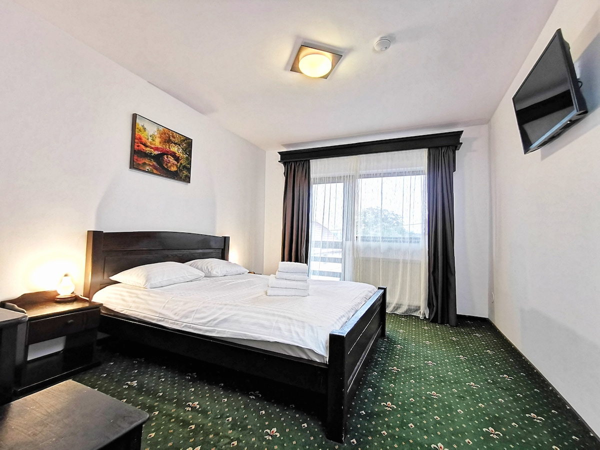 A well-appointed bedroom features a spacious bed with white linens and neatly arranged towels. A bedside table is positioned beside the bed, and a painting adds a touch of color to the wall. Light enters through large windows, draped with dark curtains.