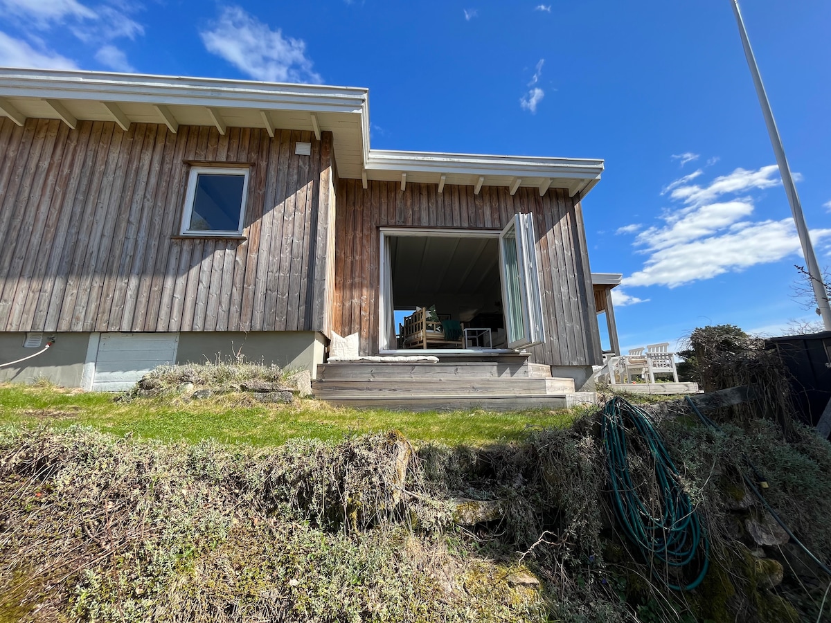The exterior of the cabin features a wooden facade and large glass doors that open onto a wooden deck. Green grass and a slight slope are visible, with blue skies and a few scattered clouds providing a bright backdrop.