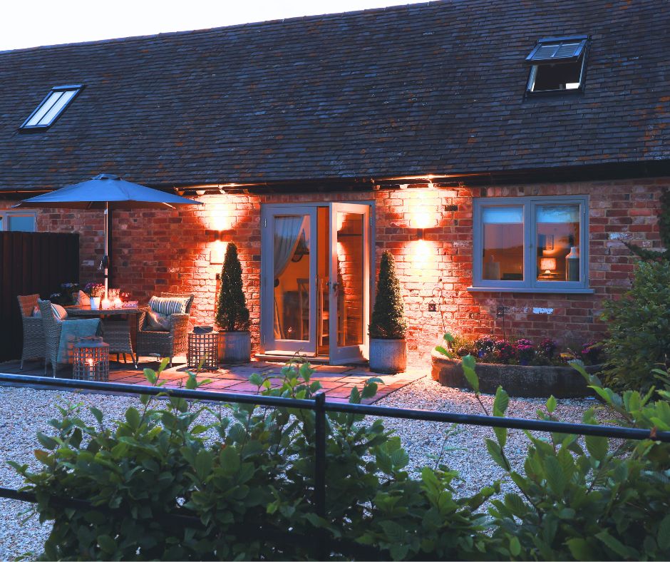 The exterior of the Piggery is softly illuminated by warm lights, highlighting the rustic brick façade. Elegant details include potted topiary, outdoor seating under a parasol, and a welcoming terrace area, all set within a peaceful garden landscape.