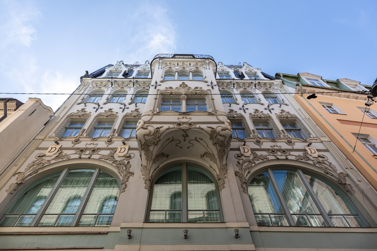 The building showcases intricate Art Nouveau architectural details, highlighted by ornate moldings and decorative elements. Large arched windows enhance the facade, allowing ample natural light. The design reflects a blend of classic elegance and historical significance under a clear blue sky.