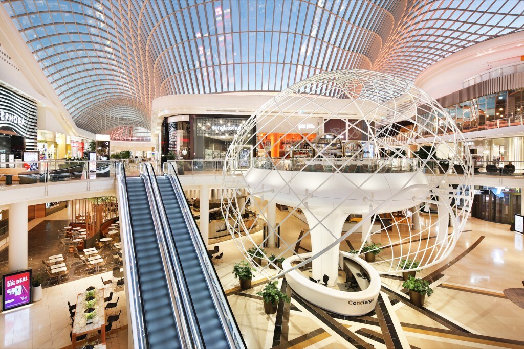 An atrium is seen featuring a modern architectural design with a large, spherical sculpture at its center. A glass ceiling allows natural light to filter in, illuminating the surrounding retail spaces and escalators. Comfortable seating areas are positioned nearby for visitors.