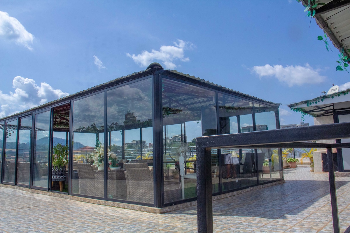 A spacious rooftop area is enclosed by large glass panels, offering panoramic views of the surrounding landscape. Enhanced with varied seating arrangements, the space features decorative plants and an inviting atmosphere created by natural light filtering through the clear glass.