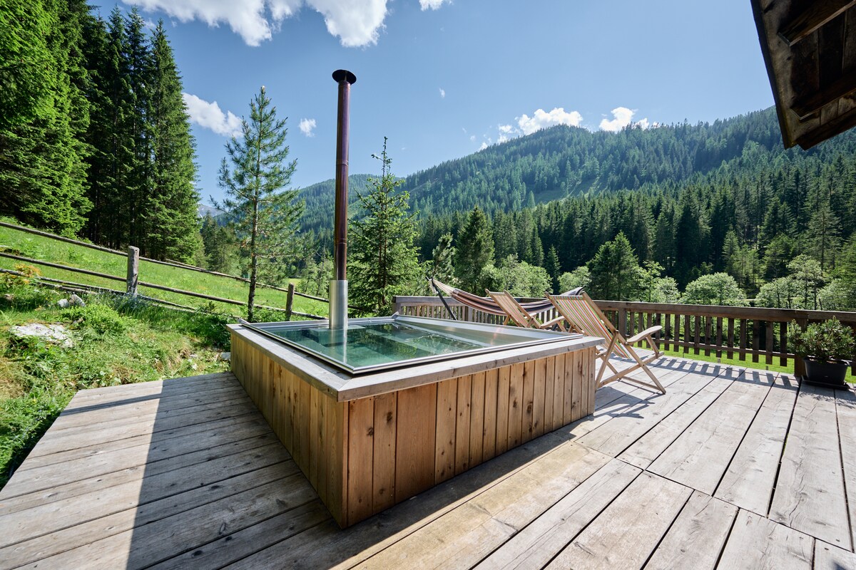 A wooden deck features a sunken hot tub surrounded by nature, with views of gentle hills and dense forest. Two lounge chairs are positioned for relaxation, offering a serene outdoor space. A chimney stands beside the tub, enhancing the rustic ambiance.