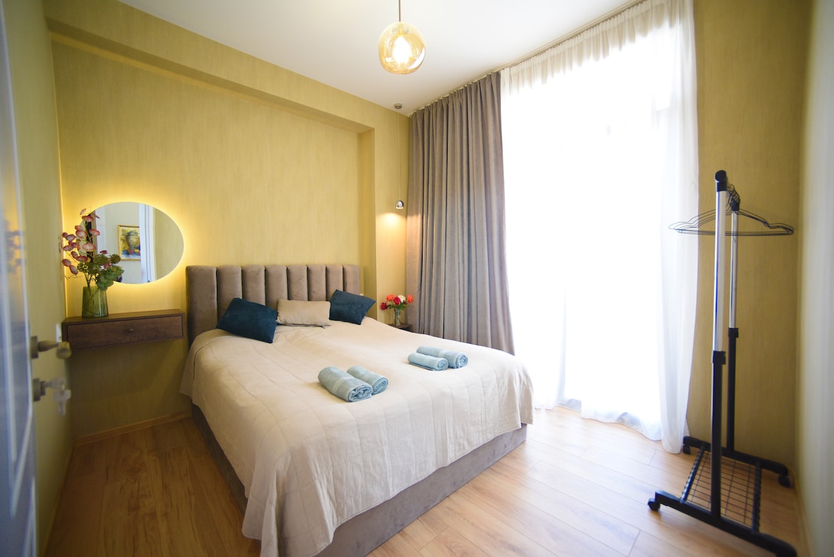 A bright bedroom features a spacious bed adorned with light bedding, flanked by two decorative pillows. Natural light streams in through the large window, complemented by soft curtains. A wall-mounted light fixture and a small table with flowers enhance the calming atmosphere. A clothing rack stands nearby.