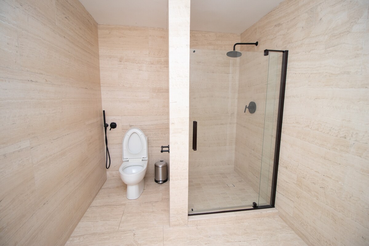 A spacious bathroom features light-colored stone walls and floor tiles. A glass shower enclosure is present, along with a contemporary toilet and a small waste bin. The shower area is equipped with a sleek black showerhead and a handheld fixture, enhancing functionality.