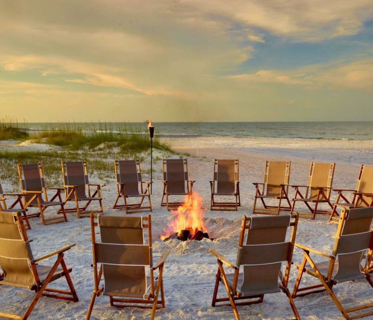 A circle of wooden beach chairs is arranged around a softly crackling campfire on the sandy shore. The tranquil ocean is visible in the background, complemented by the gentle hues of the sky during sunset. Tall grasses frame the scene, enhancing the natural setting.