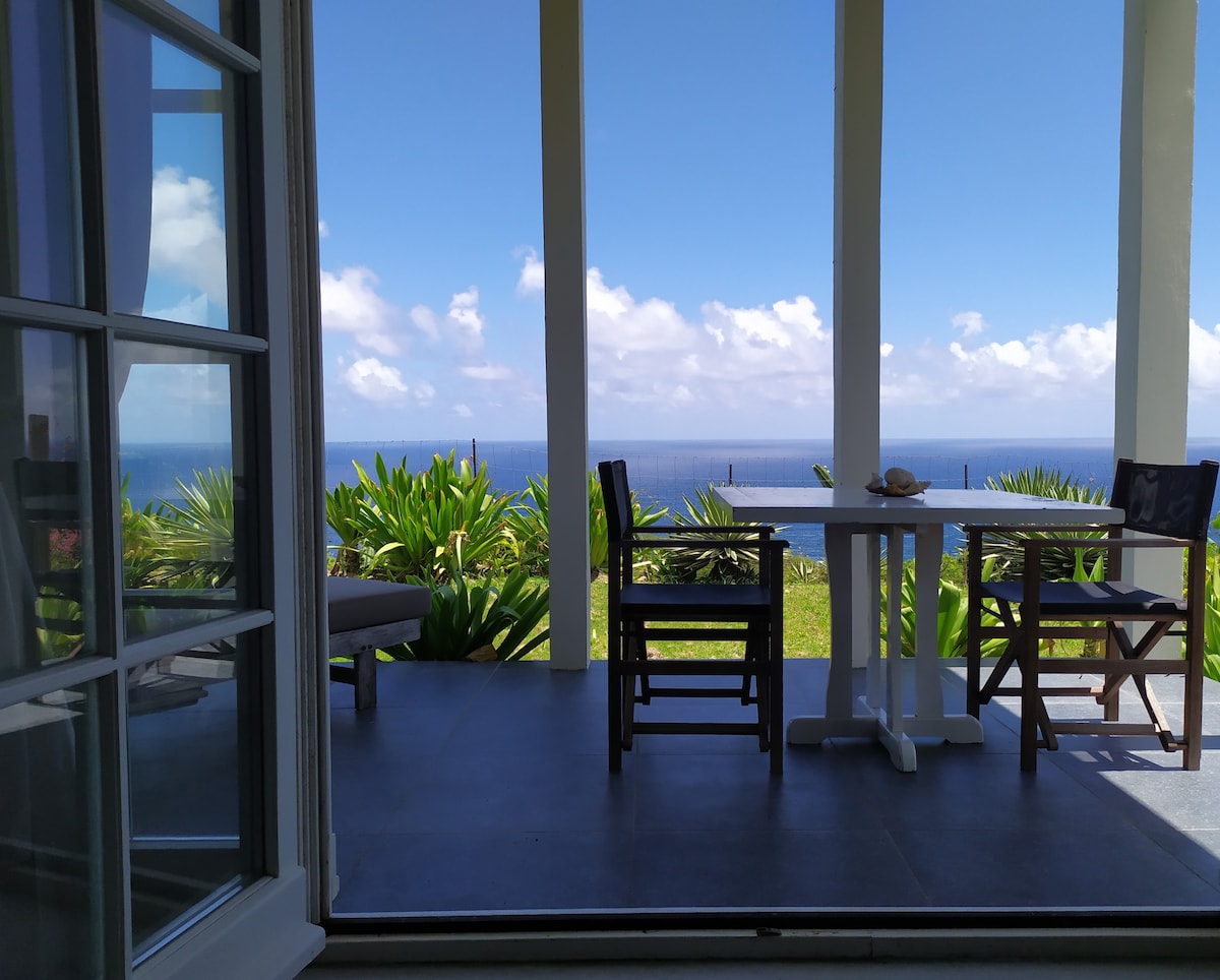 A serene outdoor seating area is visible, featuring a table and folding chairs set against a backdrop of lush greenery and ocean views. Bright blue skies and scattered clouds enhance the tranquil setting, inviting relaxation and enjoyment of the scenery.