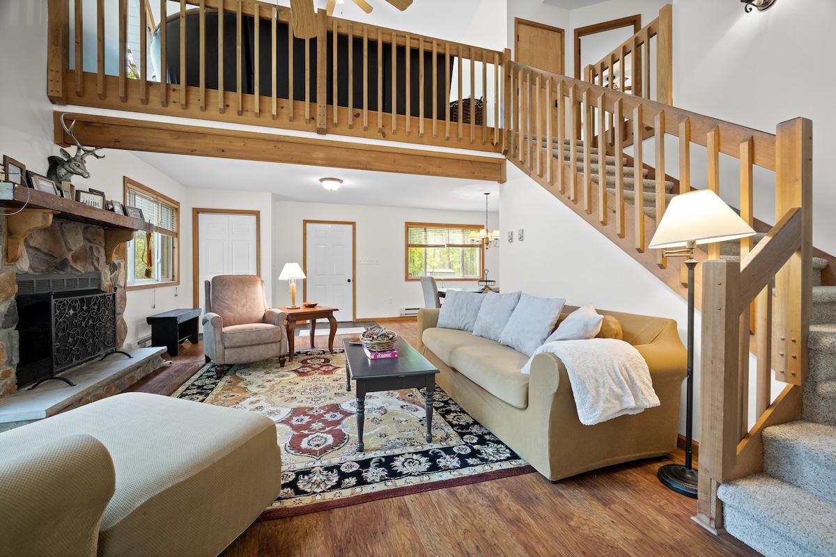 A spacious living area features a cozy seating arrangement, including a large sofa and armchair, set on a decorative area rug. Natural light fills the room through several windows. A wooden staircase leads to an upper level, while a stone fireplace adds warmth to the space.