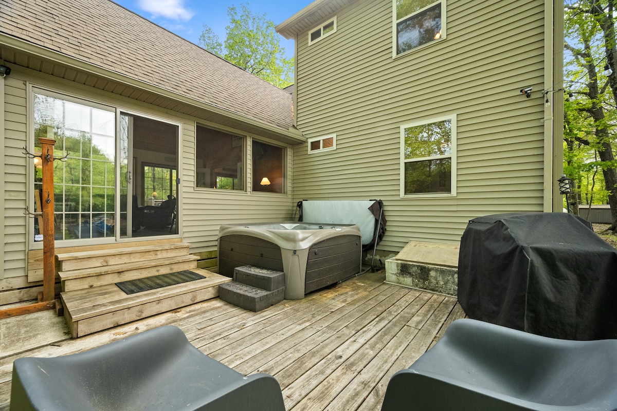 An outdoor patio area features a wooden deck with a hot tub and seating. The entrance to the home is marked by steps leading up to a sliding glass door. A grill is positioned nearby, surrounded by lush greenery and natural light.