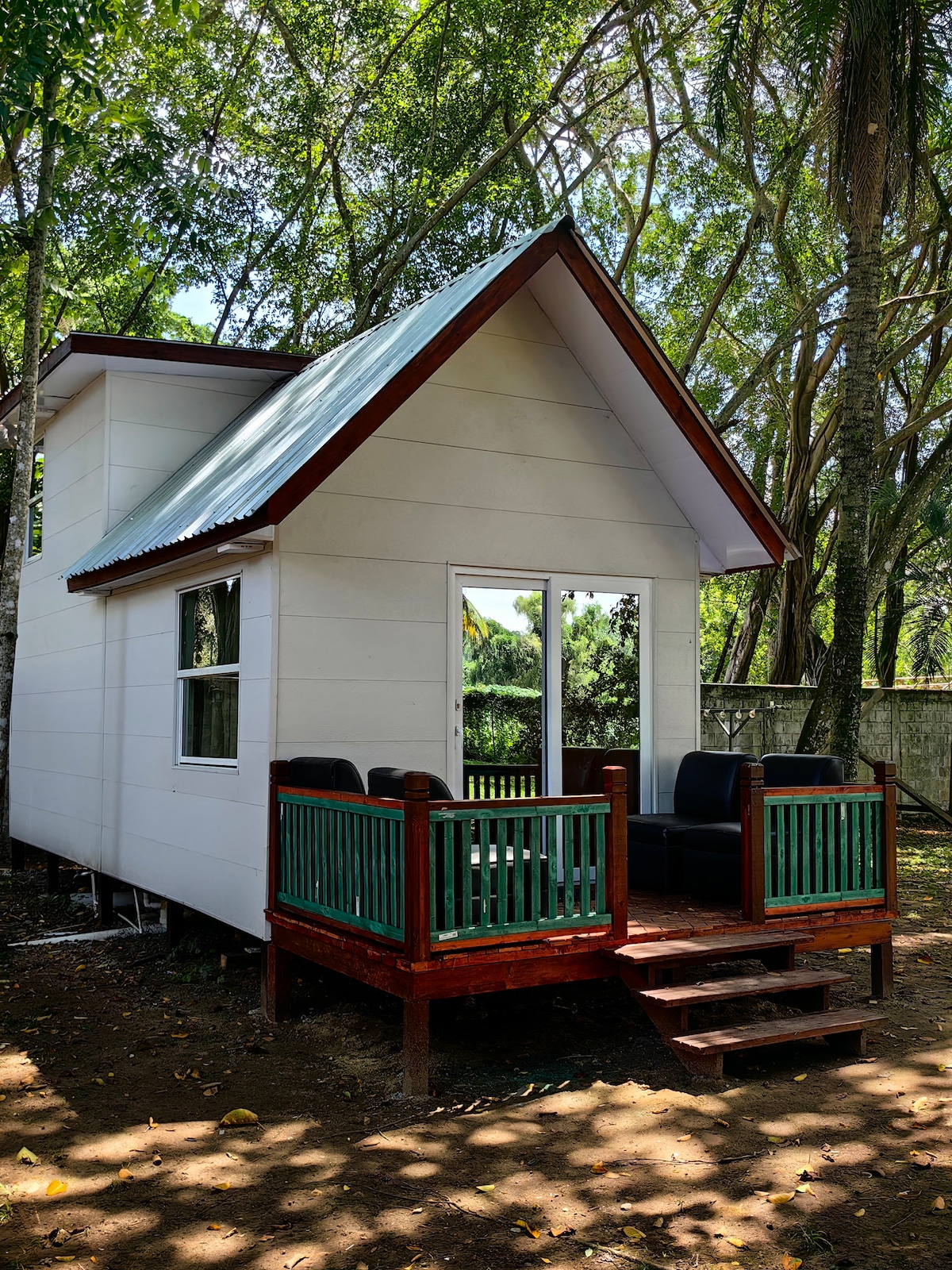 A small white house with a sloped roof sits on a spacious deck surrounded by greenery. Outdoor seating is arranged on the deck, inviting relaxation. Large windows reflect natural light, while shaded areas from surrounding trees provide a serene ambiance.