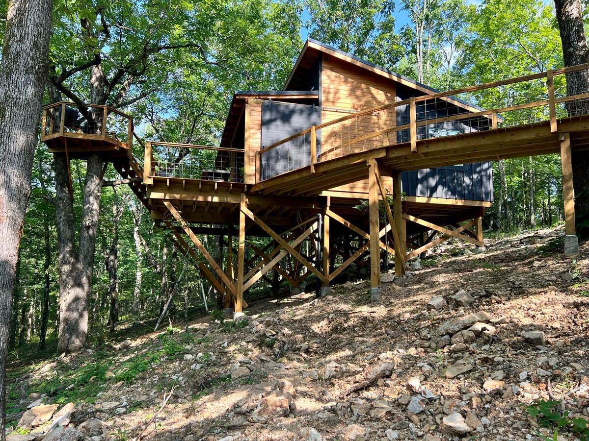 The cabin is elevated on wooden stilts, surrounded by trees. A spacious deck extends from the structure, providing outdoor accessibility. The modern design features large windows that allow for natural light, seamlessly integrating the home with the forest environment.