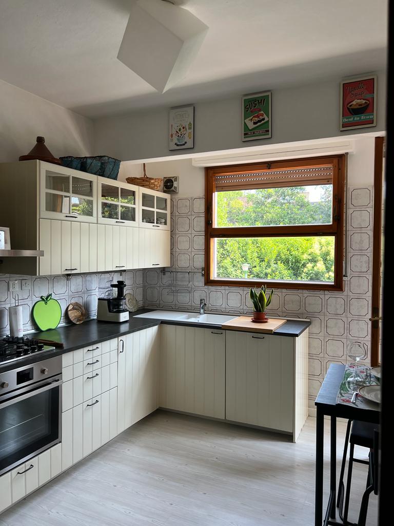 The kitchen features light cabinetry and a black countertop, equipped with a gas stove and an oven. A large window illuminates the space, providing a view of greenery outside. Shelves display decorative items, enhancing the room's functionality.