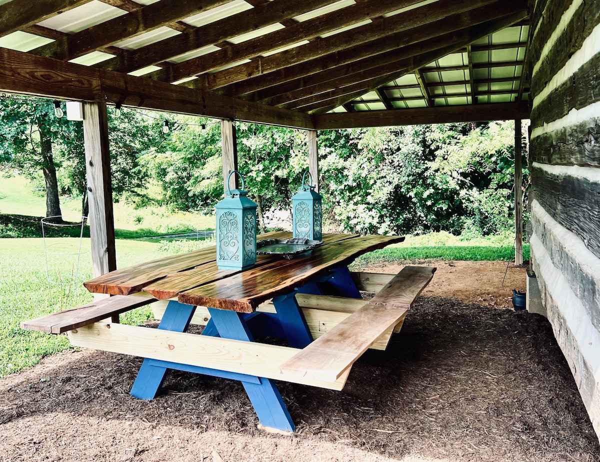 A spacious picnic table with a natural wood finish is situated under a covered porch. Two decorative lanterns are placed on the table, surrounded by grass and lush greenery. The area offers a comfortable setting for outdoor dining or relaxation.