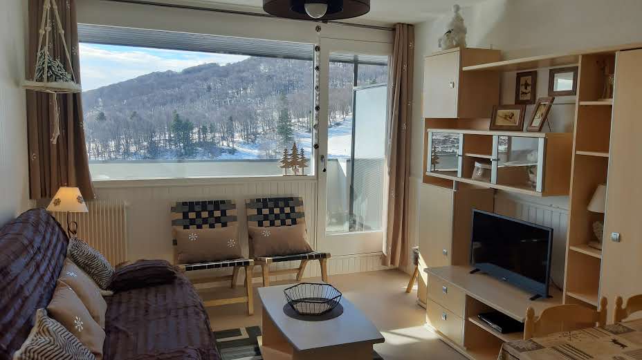 A bright living area featuring a modern design is shown, with a cozy couch and patterned chairs. A coffee table sits in front, and a TV unit is positioned alongside a large window offering views of the surrounding snowy landscape.