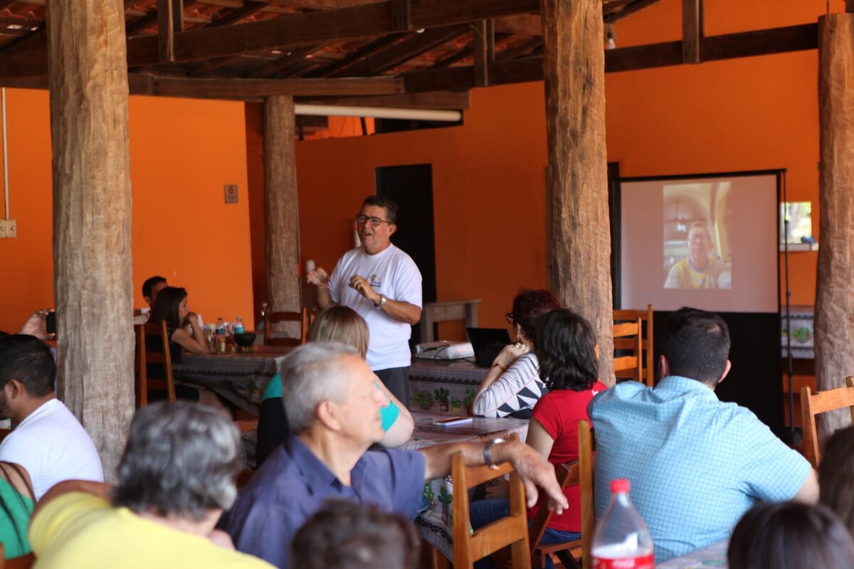 A spacious dining area is shown with wooden beams supporting the ceiling. Attendees are seated at tables with a projector screen displaying an image. The orange-painted walls create a vibrant atmosphere, while bottles of beverages are visible on the tables.