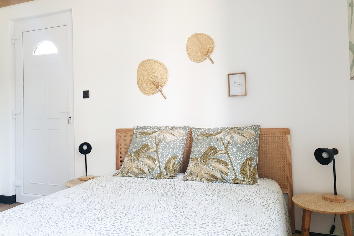 A spacious bed is presented with decorative pillows featuring a leafy design. On each side of the bed, small tables hold minimalist lamps. Two woven fans are mounted on the wall, complementing the light-colored, neutral decor. A door is visible in the background.