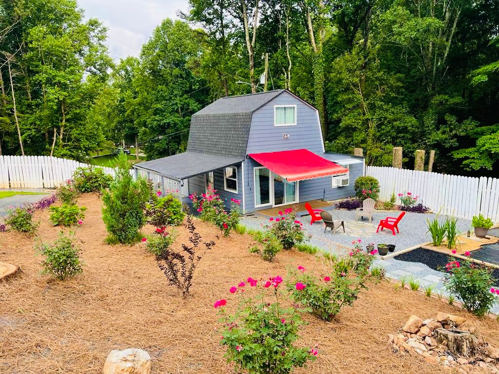 The exterior of a two-story guesthouse is set amidst natural greenery, surrounded by a landscaped garden featuring colorful flowers. A red awning shades the entrance, while a gravel path leads to seated areas. The homey structure exhibits a blend of rustic charm and modern design.