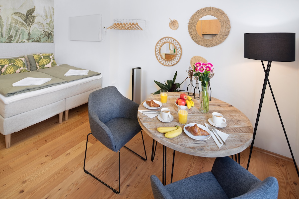 A dining area is set with a round wooden table adorned with breakfast items, including fruit, pastries, and beverages. Two stylish chairs complement the table. In the background, a cozy sleeping area is visible with a neatly arranged bed, decorated with tropical-themed cushions and artwork.