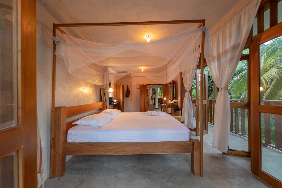 A spacious room features a four-poster bed with a light canopy, complemented by wood accents and soft lighting. Large windows allow natural light to fill the space, while sheer curtains create a gentle flow. A balcony is visible, enhancing the connection to nature.