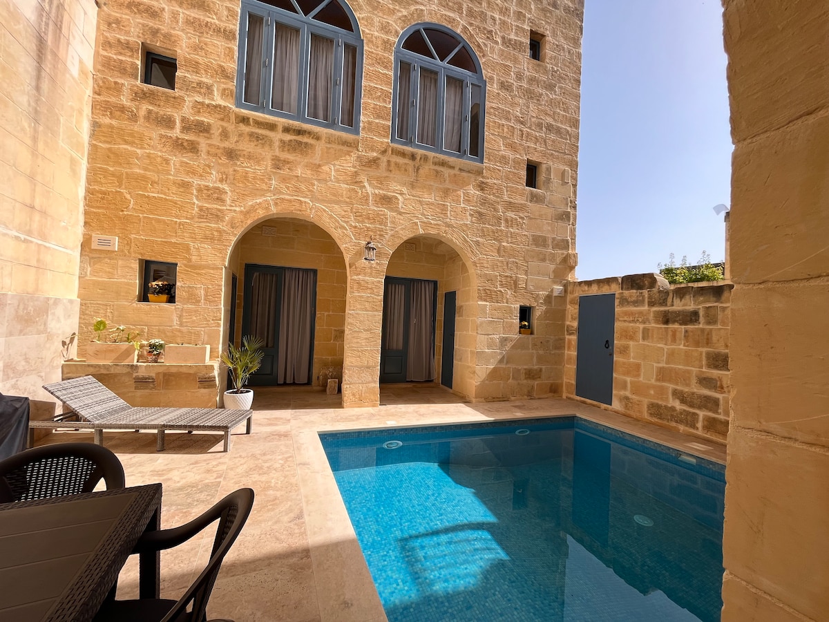 A private outdoor pool glistens under the sun, framed by natural stone walls. Sun loungers are positioned nearby, creating a relaxing space. Large windows with blue shutters provide access to the adjacent rooms, enhancing the inviting atmosphere of the courtyard.