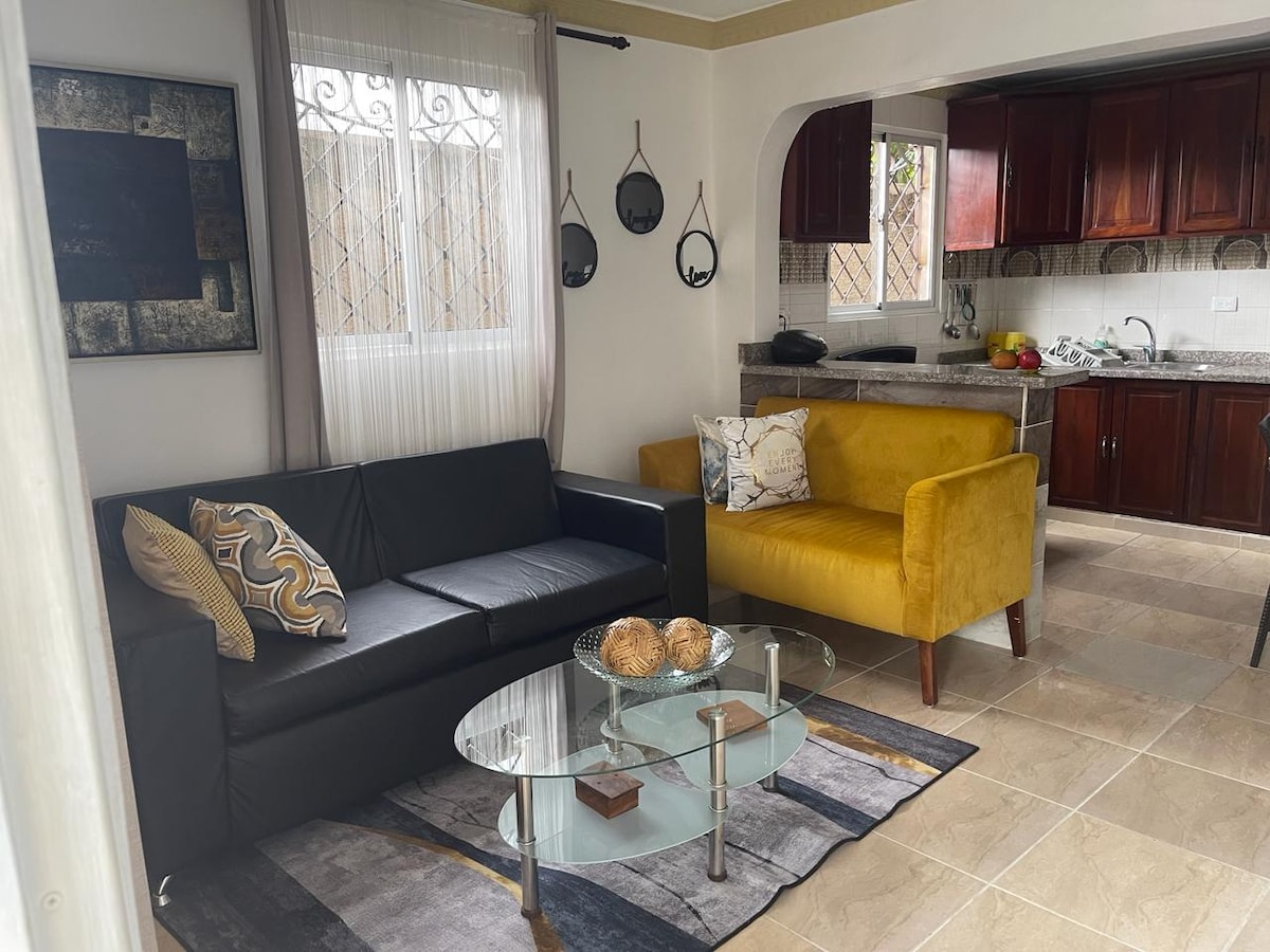 A living area features a black sofa and a yellow accent chair, positioned around a glass coffee table. A patterned area rug adds texture, while mounted wall art and a large window bring light to the space. The adjoining kitchen is visible in the background.