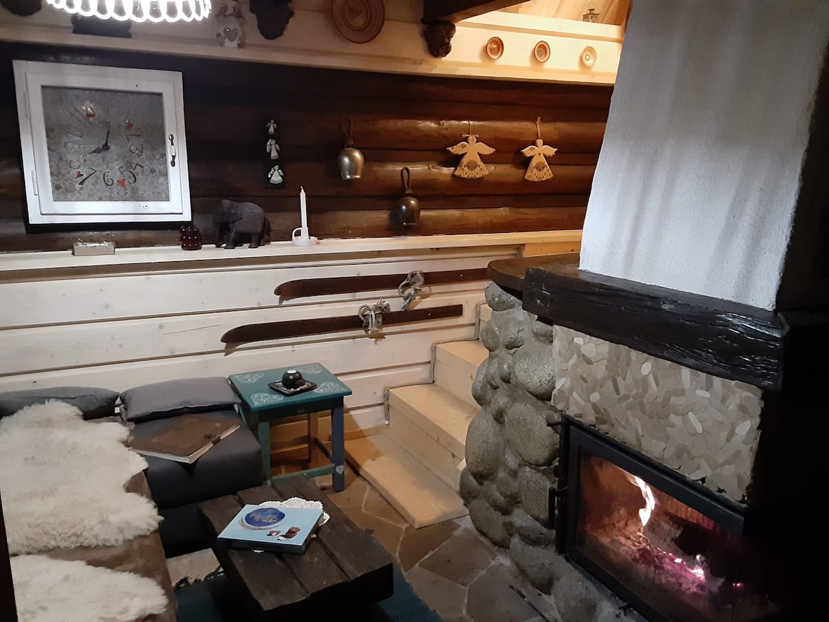 A rustic living room features wooden walls and a welcoming fireplace. A cozy sofa is positioned near a stone fireplace, and wooden skis are displayed on the wall. A coffee table with a book and small decorative items adds to the homely ambiance.