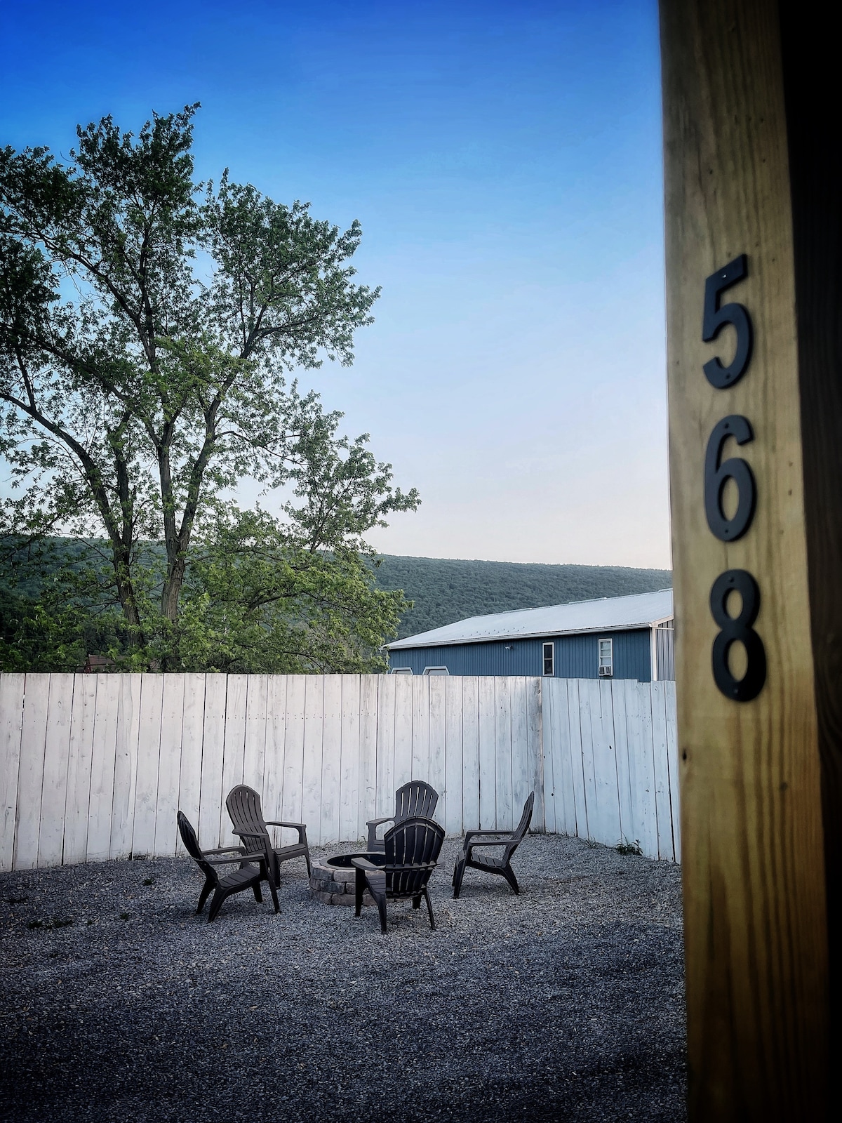 An outdoor seating area is visible, featuring four dark wooden chairs arranged around a small circular table. A white fence encloses the gravel space, with a tree in the background and hills visible beyond.
