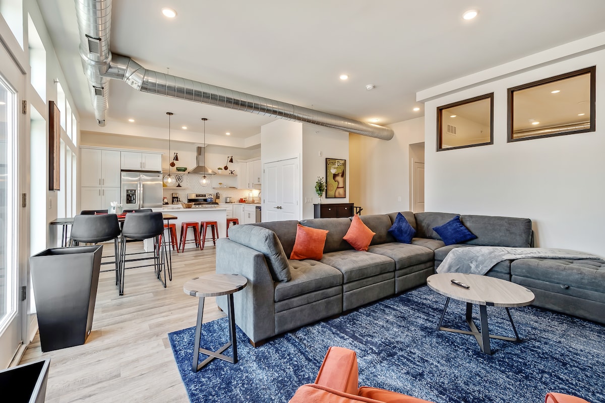 An open-concept living area reveals a spacious sectional sofa adorned with decorative cushions. Soft lighting highlights modern fixtures in the kitchen, where bar stools are arranged around the counter. Large windows invite natural light, enhancing the airy atmosphere of the room.