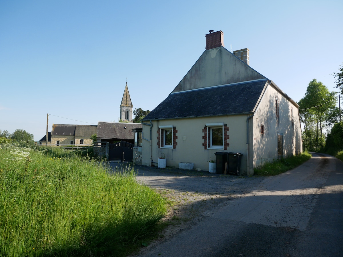 A quaint house is situated along a gravel road, flanked by lush greenery. The building features a pale exterior with red window accents. In the background, the steeple of a church rises above the landscape, providing a sense of local history and charm.