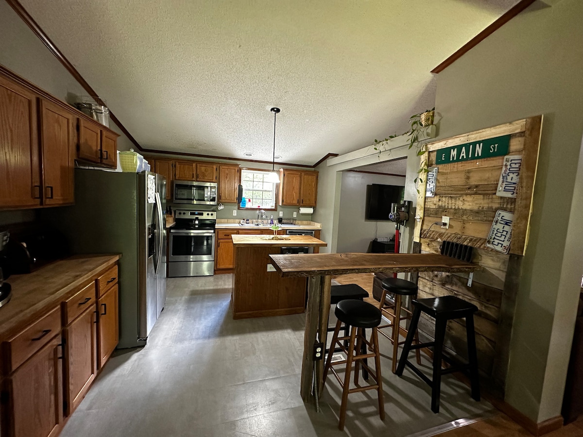 The kitchen features wooden cabinets and stainless steel appliances, including a refrigerator and an oven. A large island provides ample countertop space. A rustic wooden bar with stools offers additional seating. Natural light fills the room through the window above the sink.
