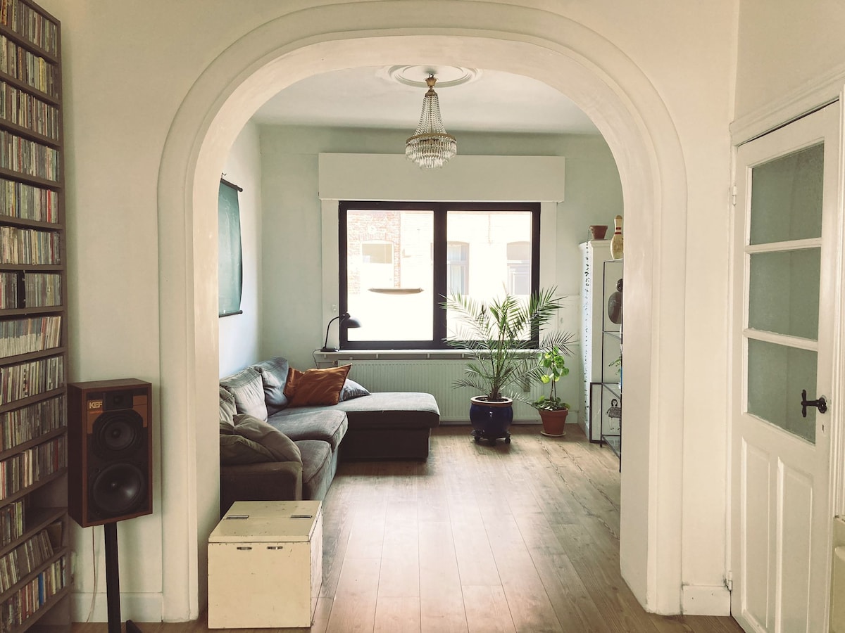 A living area is framed by an elegant archway, featuring a comfortable sectional couch and decorative indoor plants. Large windows provide natural light, with a small table placed beside a speaker. Wooden flooring enhances the spacious feel of the room.