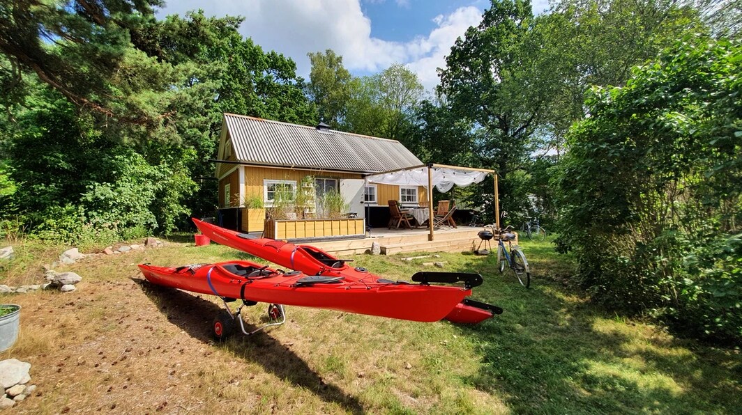 Two red kayaks are positioned on grassy ground in front of a wooden cabin surrounded by lush greenery. The cabin features a covered outdoor seating area and large windows. A bicycle is parked nearby, adding to the outdoor recreational atmosphere.