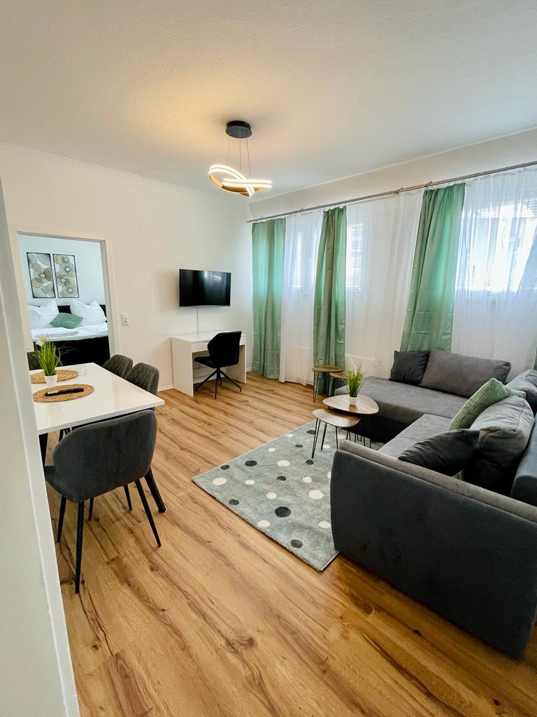 A modern living area showcases a comfortable sectional sofa in gray, paired with a light area rug featuring subtle patterns. A sleek dining table accommodates several chairs, and a wall-mounted television is positioned across from the seating. Natural light filters through sheer and solid curtains.