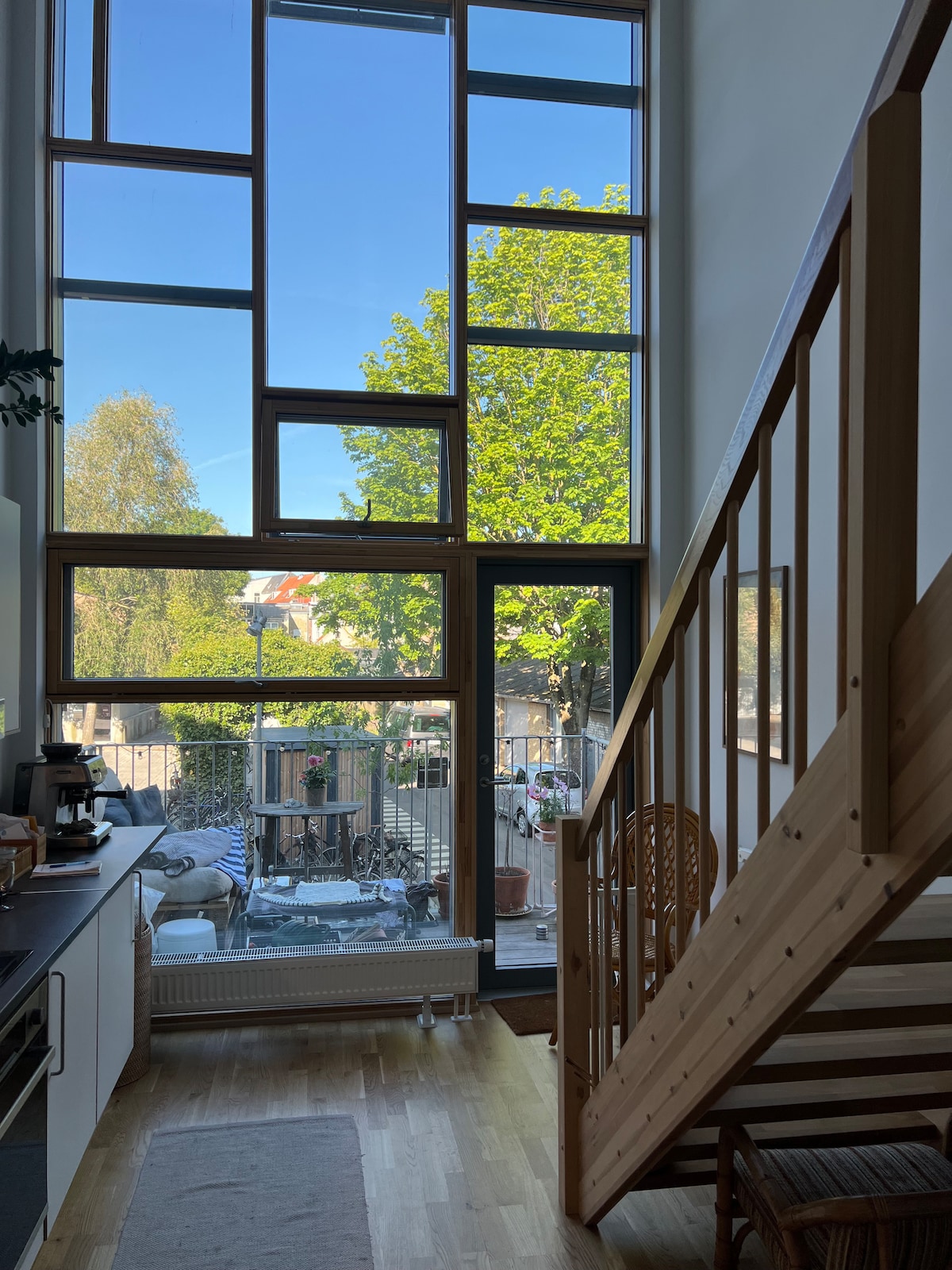 Large windows allow ample natural light to fill the space, offering views of outdoor greenery. A staircase is visible on the right, and the kitchen area features modern appliances. A cozy outdoor seating area can be seen through the open door.