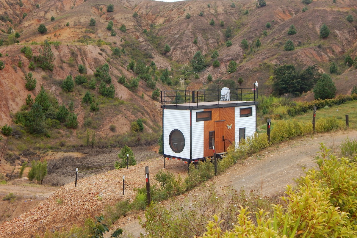The tiny house is situated on a hillside, surrounded by natural terrain. A dirt path leads to the elevated structure, which features round windows and a terrace. Green plants and shrubs frame the house, providing a sense of integration with the landscape.