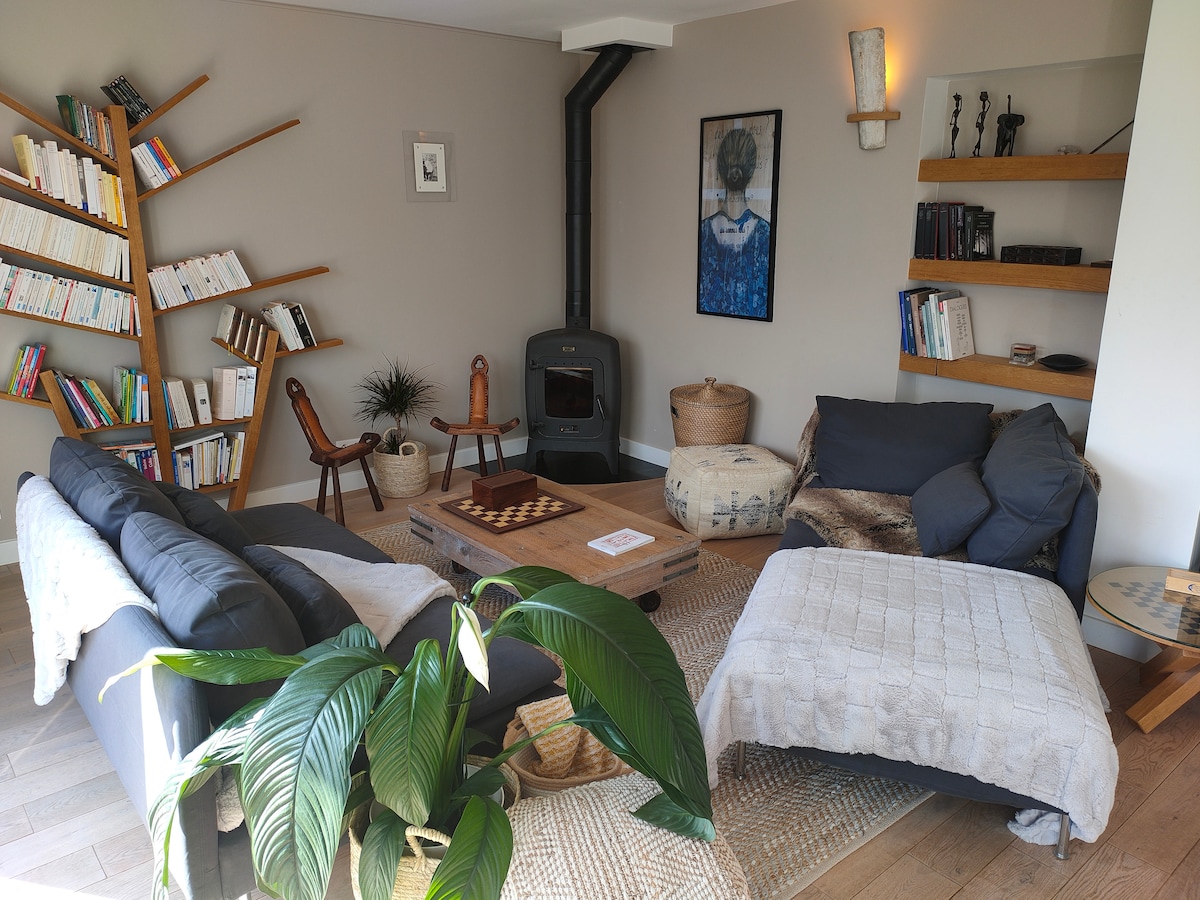 A cozy living area features a combination of dark-colored sofas and a comfortable lounge chair, arranged around a wooden coffee table. A bookshelf filled with various books is situated against one wall, complemented by decorative items on shelves and a potted plant adding a natural touch.