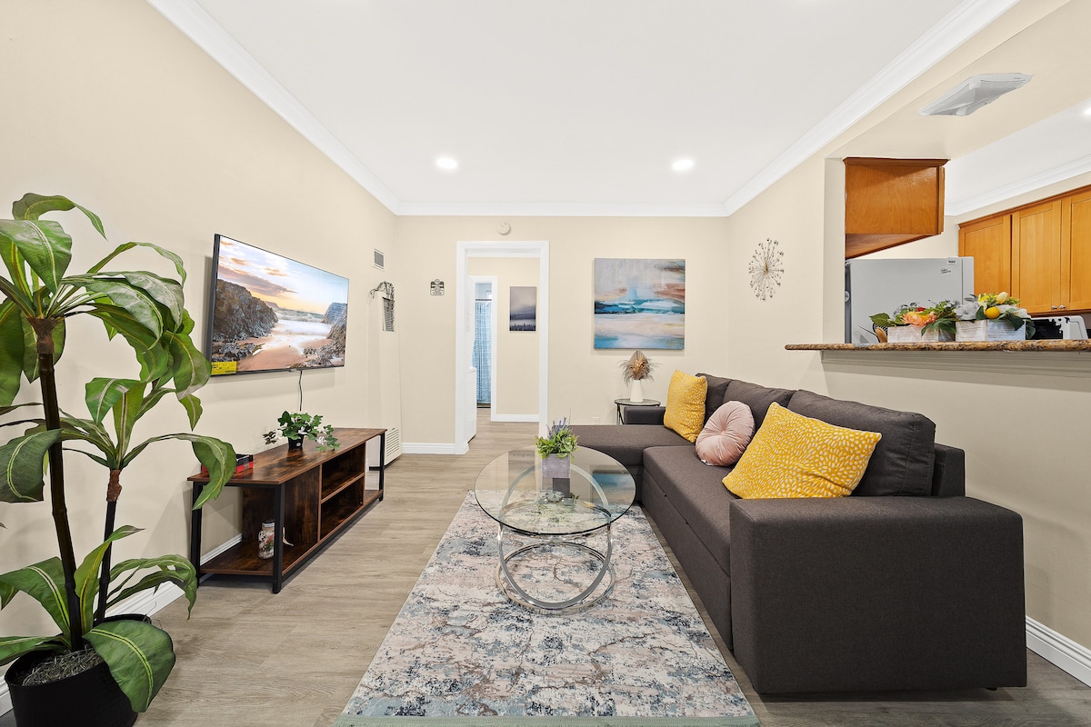 An open concept living area features a comfortable dark sofa adorned with colorful pillows. A glass coffee table sits on a textured rug, complemented by a large plant in the corner. The wall-mounted TV displays a scenic image, with a light-colored kitchen and hallway visible in the background.