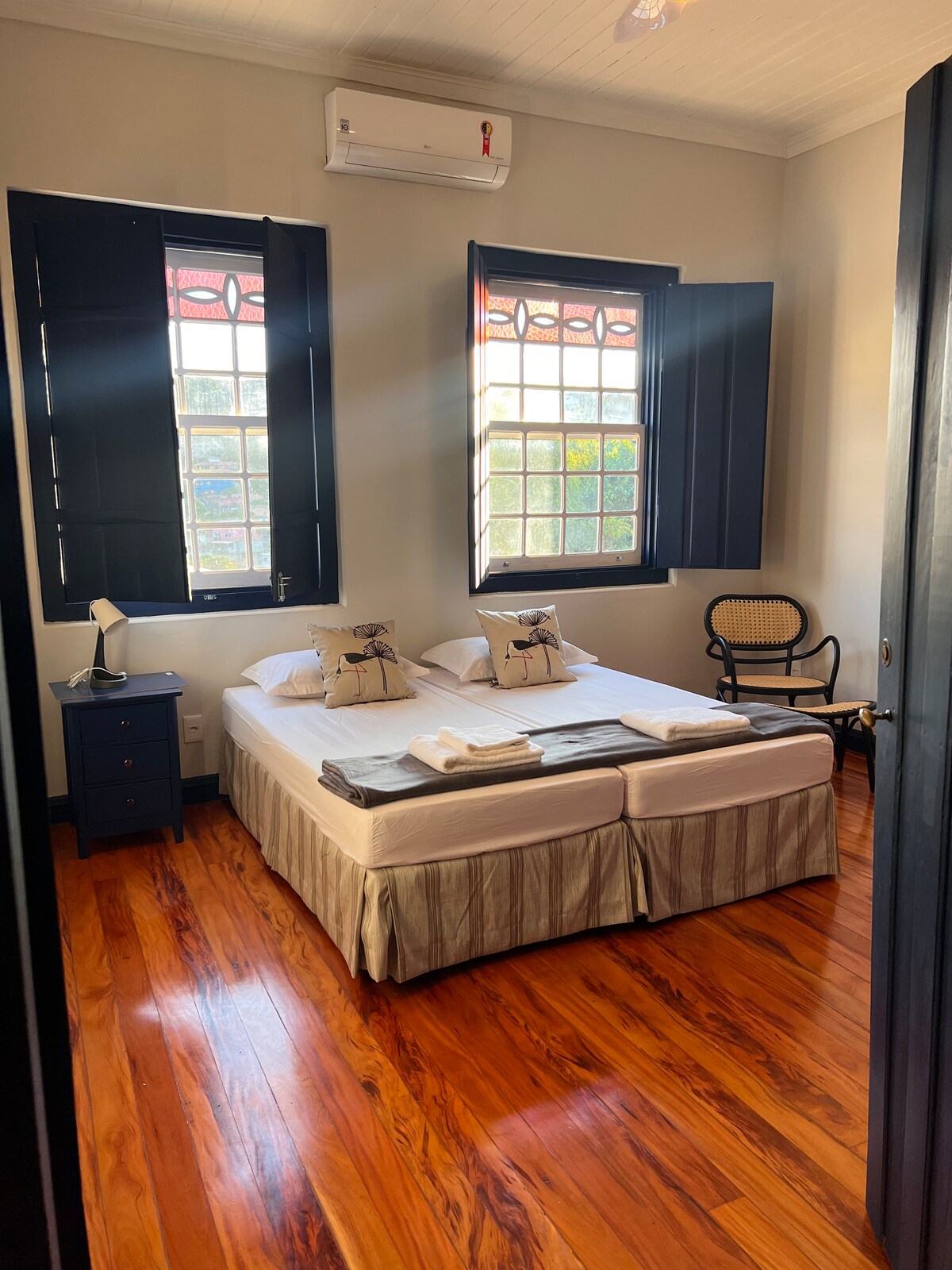 A well-appointed bedroom is presented with two spacious beds, complemented by fresh linens. Natural light filters through two large, opened windows, enhancing the warm wood flooring. A bedside table and a stylish chair are included, along with an air conditioning unit mounted above.